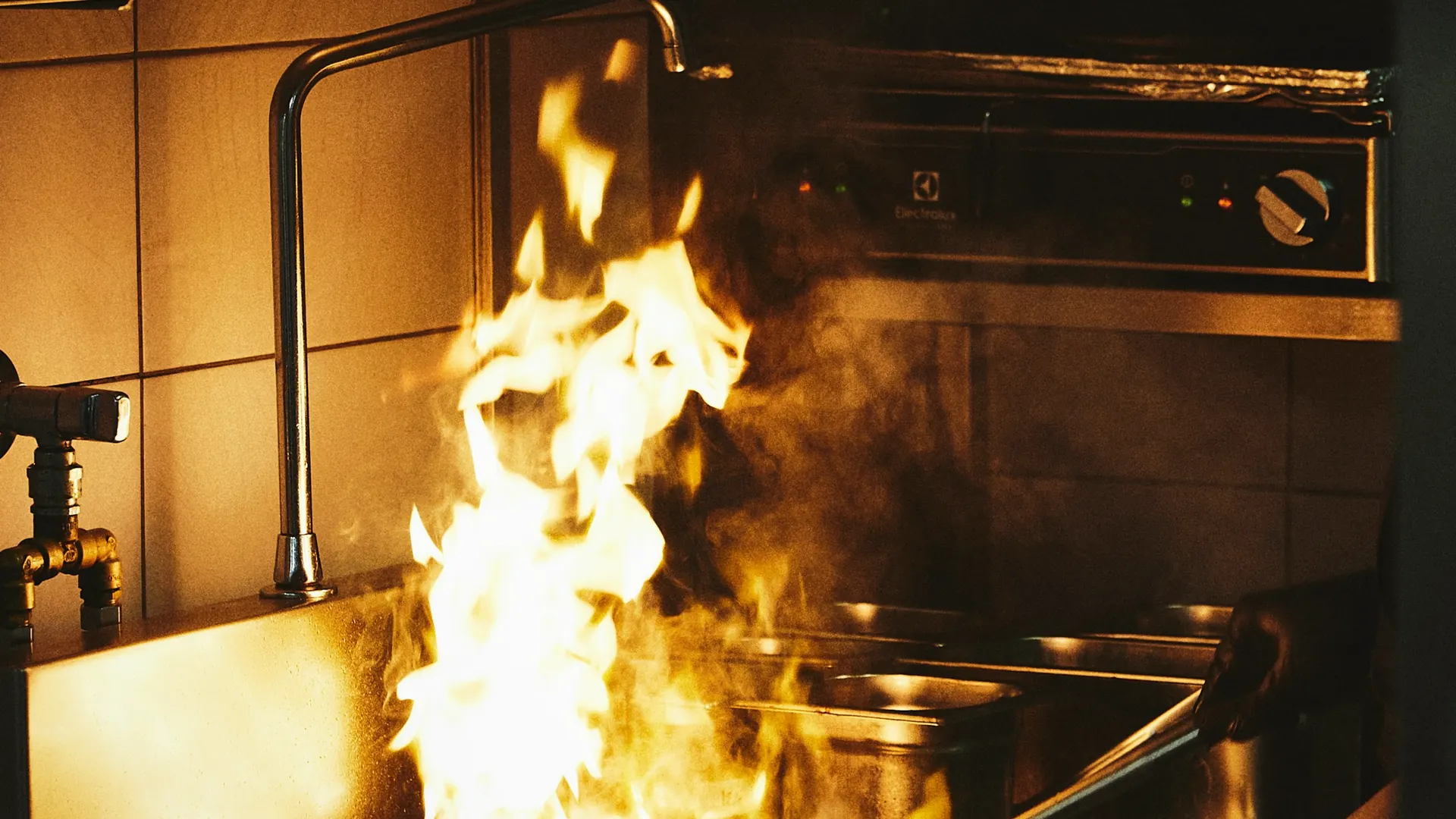 Handen van een kok boven een pan met vlam in de open keuken, met groene tegels op de achtergrond